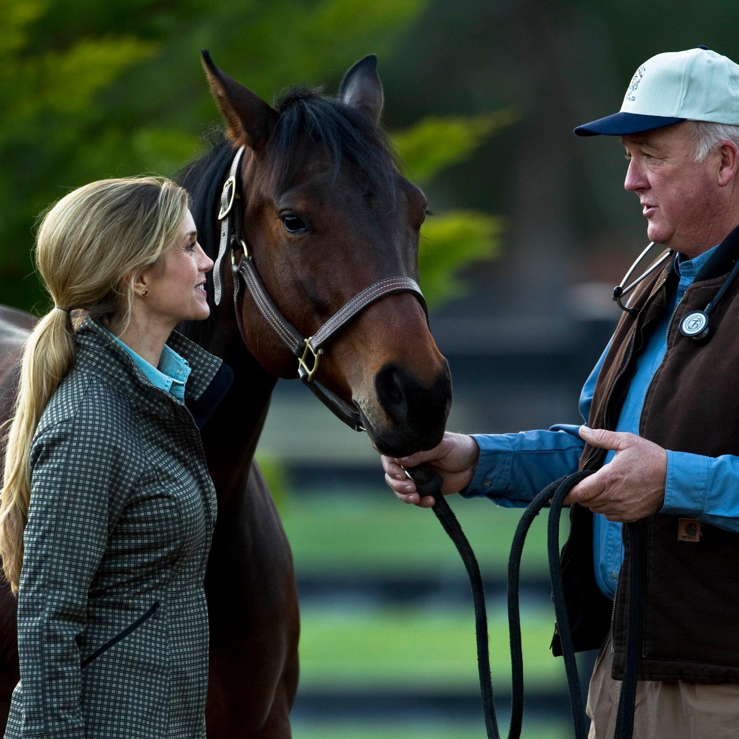 A gentleman with a white cap talking with a blond haired smiling woman, with a dark chestnut colored horse standing waiting to be ridden.